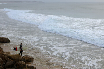 a lone surfer carrying his surfboard along the rocky coast looking for an opportunity to enter the waves and join the surf on a rainy early morning surf, great ocean road, Victoria, Australia