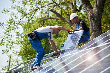 Team of technicians mounting heavy solar photo voltaic panel on tall steel platform on green tree and blue sky background. Exterior solar panel voltaic system installation, dangerous job concept.