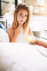 Obraz na płótnie Canvas Attractive young female holding glass of orange juice and looking at camera while sitting in comfortable restaurant, beautiful blonde hair woman enjoying rest during work break in modern interior