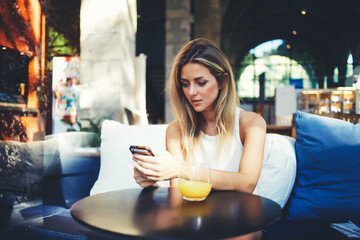 Portrait of a cute blonde female read something on her smart phone while sitting in modern coffee shop, beautiful young hipster girl using cell telephone while enjoying her free time in expensive cafe