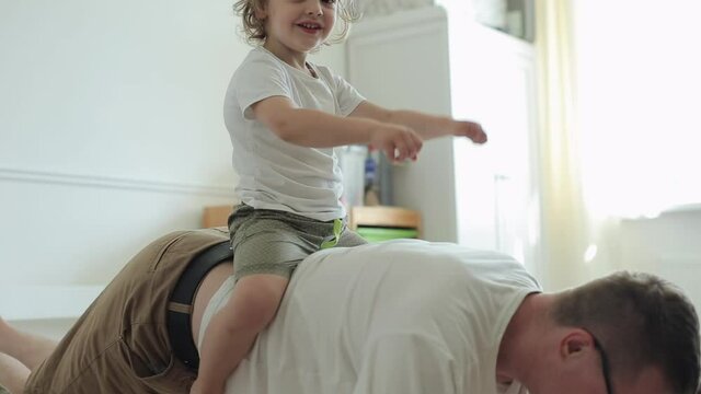 A little blond boy with big brown eyes sits on his father's back imagining that this is a horse while his father does an exercise and push-up off the floor. Close-up
