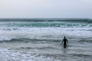 an unidentfiiable lone male surfer leading his surfboard through the waves off the beach on a rainy morning, as his friends surf in the background, Bell's beach, great ocean road, Victoria, Australia