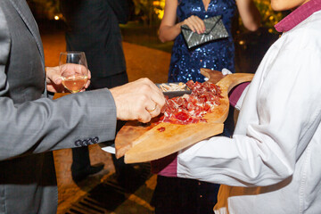 La mano de una persona coge una loncha de jamón que le ofrece un camarero en una bandeja de madera - obrazy, fototapety, plakaty