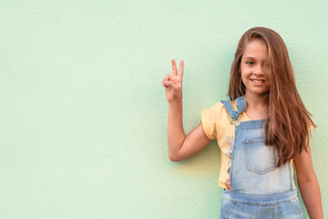 Portrait of a little girl child in a denim jumpsuit 9-13 years old on an empty wall, the girl shows two fingers symbolizing that everything is good