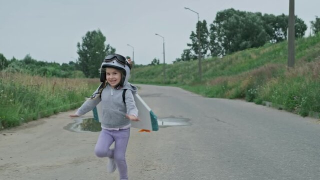 Little girl having fun with white helmet and jetpack outdoors in slow motion. Imagination and freedom. Cute little girl running on rural road acting to be pilot