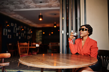 Portrait of african american woman, retro hairstyle with eyeglasses, wear orange jacket posing at restaurant with glass of wine.