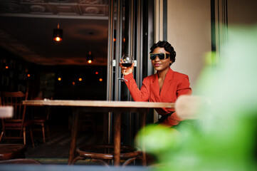 Portrait of african american woman, retro hairstyle with eyeglasses, wear orange jacket posing at restaurant with glass of wine.