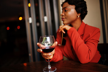 Portrait of african american woman, retro hairstyle with eyeglasses, wear orange jacket posing at restaurant with glass of wine.