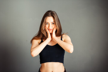 Obraz premium A girl in a black top with her hair loose with hands on her cheeks is smiling on a gray background. Without makeup, without retouching.