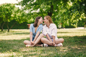 Fototapeta premium Two beautiful girls have fun in the park on a sunny summer day. The sisters are sitting on a green lawn, laughing.