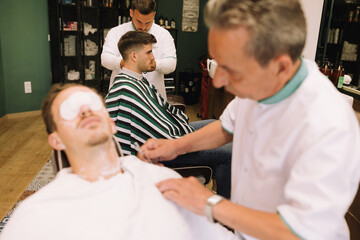 barber cutting the beard of an attractive young brown man with cotton wool in his eyes