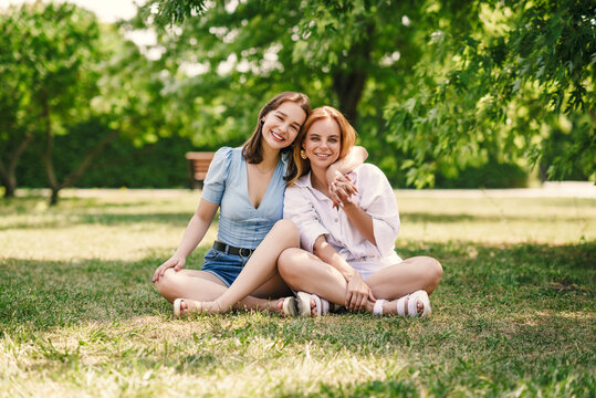 Two beautiful girls have fun in the park on a sunny summer day. The sisters are sitting on a green lawn, laughing.