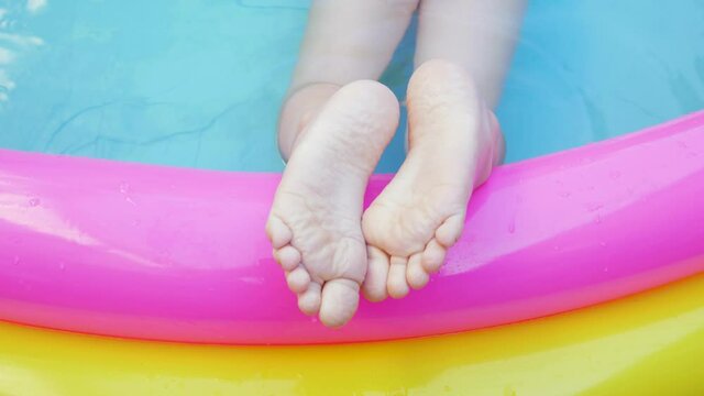 Kids feet laying on colorful rainbow inflatable swimming pool. Young girl in pink swimsuit playing in water on a backyard. Summer relax, active lifestyle, children swim fun home leisure,hot weather 4K