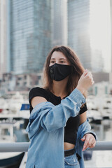 Young european woman with brown hair wearing denim shirt and protective face mask with filter. Female standing at city street during Covid 19 pandemic