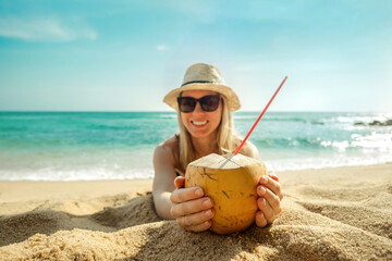Happy Young Tourist Smiling Caucasian Woman in hat with coconut in her hands on beach at sunny day. Beautiful Sunset light on coastline.

