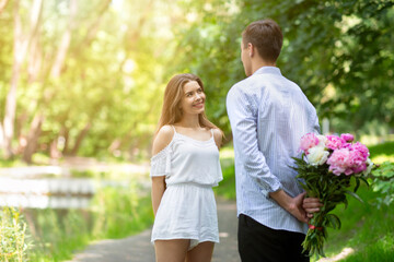 Fototapeta premium Young man congratulating girlfriend with birthday, hiding flower bouquet behind his back at park. Empty space