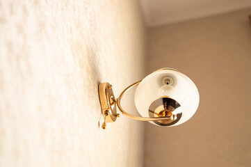 a wall lamp with a white round plafond with a gold holder and frame is attached to the wall of the apartment corridor on a uniform background taken from the lower point of the shooting with natural li