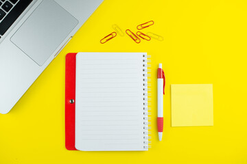 Red notepad, pen, colorful clips and laptop on yellow background. Flat lay with copy space. Workplace in the office.