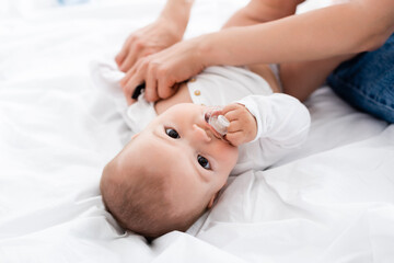 selective focus of mother wearing baby romper on infant son with pacifier looking at camera