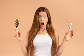Surprised millennial girl with long straight hair holding scissors and brush on beige background