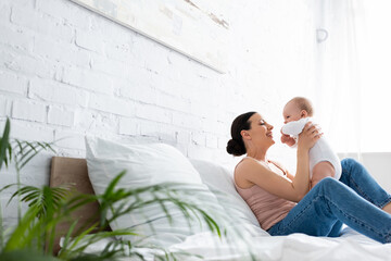 selective focus of happy mother sitting on bed and holding in arms cute infant son