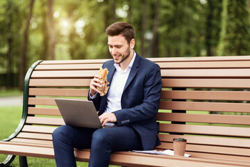 Millennial businessman eating sandwich and working on laptop during lunch break at park