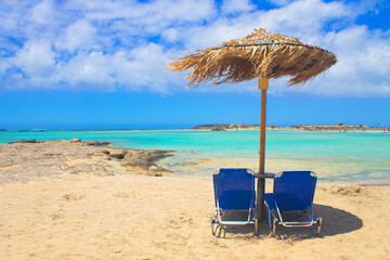 Two blue sun beds and umbrella on pink sand beach with rocks, turquoise water and clouds in blue sky.