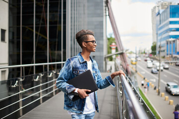 Positive hindu students holding modern laptop computer in hands enjoying leisure time in downtown admiring metropolis scenery.Cheerful hipster guy with netbook walking in urban setting in free time