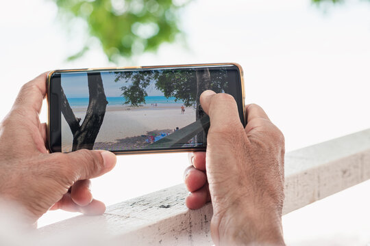 Close up hands of man holding mobile phone open a picture of sea beach after he take a photo by his smart phone.He spends his holidays with natural travel. He touched the screen to see his craft work.
