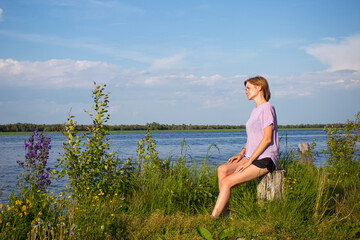 Naklejka na ściany i meble The concept of walks alone. Young girl with short hair in purple T-shirt and shorts sits on river bank. Summer sunny evening. Copyspace. Cozy cute rural landscape.
