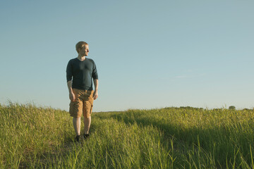 The concept of walking alone. A young Caucasian blond man walks along a path in a meadow on a sunny day. A cozy look in calm warm colors.