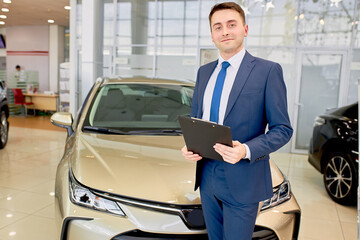 affable caucasian young consultant ready to serve and help customers in cars dealership, man is suit stand next to new car and look at camera