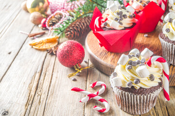Christmas cupcakes. Xmas hot chocolate cupcake with peppermint candy cane, chocolate drops and marshmallow. Wooden background with Christmas decor and fir tree branches. Sweet Christmas baking/