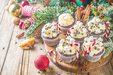 Christmas cupcakes. Xmas hot chocolate cupcake with peppermint candy cane, chocolate drops and marshmallow. Wooden background with Christmas decor and fir tree branches. Sweet Christmas baking/
