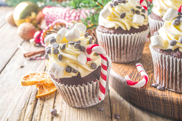 Christmas cupcakes. Xmas hot chocolate cupcake with peppermint candy cane, chocolate drops and marshmallow. Wooden background with Christmas decor and fir tree branches. Sweet Christmas baking/