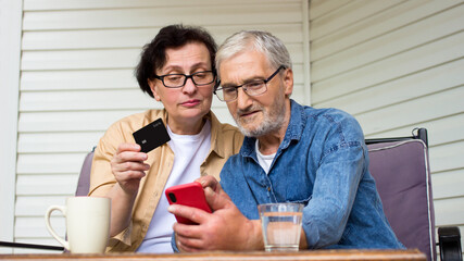 Retirement senior couple shopping online using smartphone and bank credit card,sitting on sofa at home.Happy 60s elderly spouses learning payment system,choosing service, product in internet store
