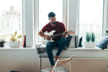 Handsome young man in casual clothing playing guitar while sitting on the window sill