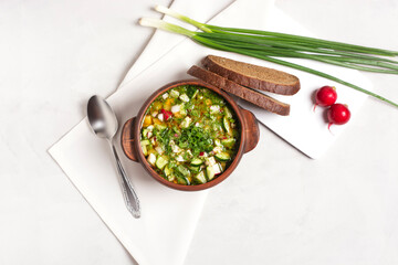 Cold okroshka with fresh vegetables and meat in a clay cup stands on a gray background, next to it...