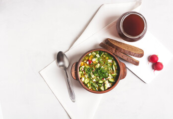 Russian okroshka with bread kvass and vegetables in a clay cup on a gray background, next to it...