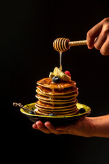 Woman pouring honey on tasty pancakes with berries on table, closeup. cooking by chef hand on dark background