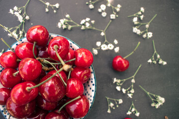 Red cherries in a bowl on a black background. Lower left corner, background decorated with small gypsophila flowers