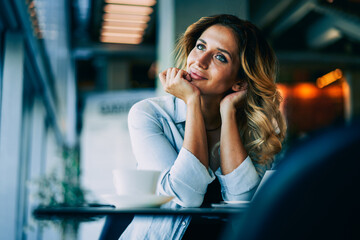 Young happy hipster girl dressed in trendy outfit dreaming while sitting at table in coffee shop.Gorgeous pondering female blogger thinking about new ideas for content in personal website indoors
