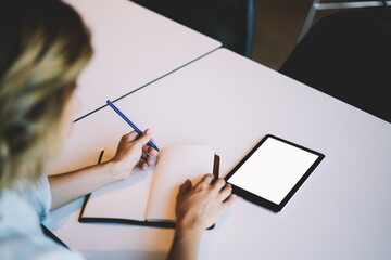 Cropped back view image of creative business woman searching information in digital tablet connected to internet with blank screen for your advertising message and recording main details to notepad