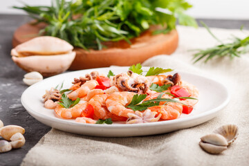 Boiled shrimps or prawns and small octopuses on white ceramic plate on a black concrete background and linen textile. side view, close up.