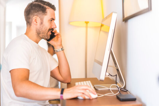 A Young Caucasian Man Telecommuting At Home On The Computer, A New Normal After The Coronavirus Pandemic, Covid-19. Work Video Call. Talking On The Phone With A Client