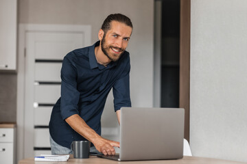A man stands near a laptop and looks into the camera with a smile, a guy works at a computer at home
