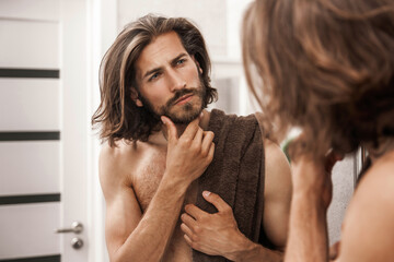 Handsome man with long hair looks at himself in the mirror after a shower with interest and touches...