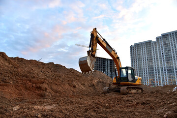Obraz premium Excavator during earthmoving at construction site on sunset background. Сonstruction machinery for excavating. Tower cranes lifting a concrete bucket for pouring of concrete into formwork