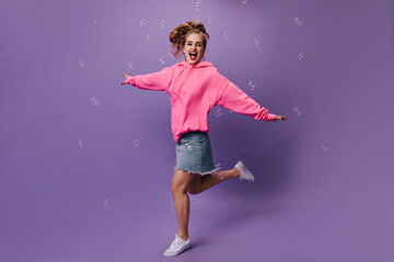 Cheerful woman in pink outfit jumping on purple background with bubbles. Full-lenght portrait of lady in loose hoodie and denim skirt running on isolated
