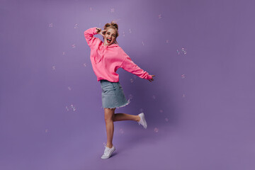 Fototapeta na wymiar Beautiful teen girl in pink hoodie showing peace sign and jumping on purple background with bubbles. Happy woman in denim skirt dancing on isolated